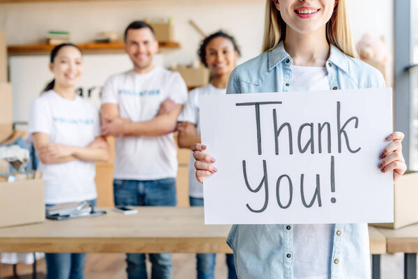 частичный вид женщины с плакатом с благодарственной надписью, стоя рядом с молодыми улыбающимися волонтерами
