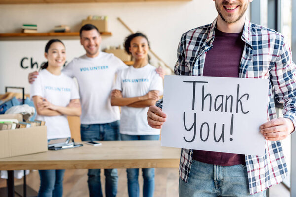 частичный взгляд человека, держащего карточку с благодарственным текстом, стоя рядом с мультикультурными волонтерами в благотворительном центре
