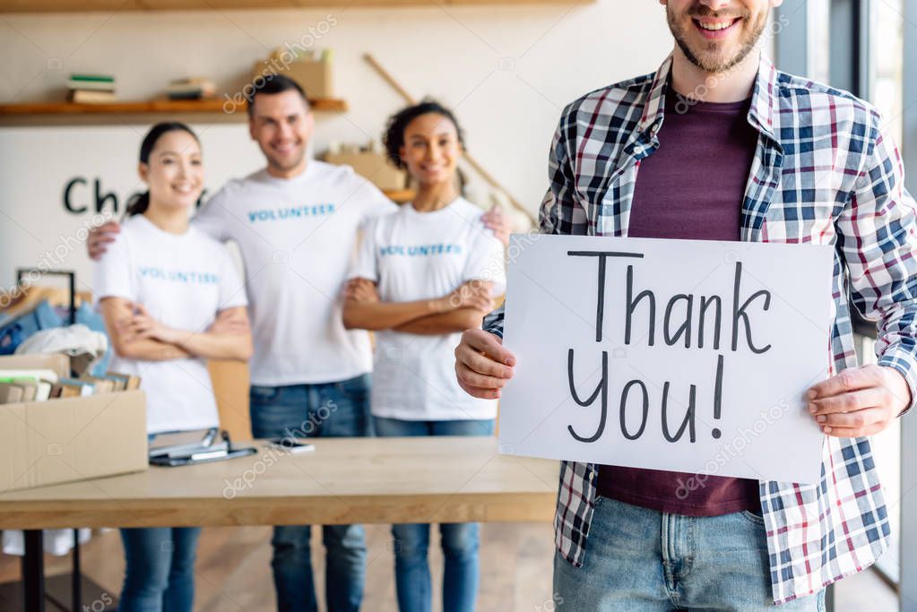 partial view of man holding card with thank you text while standing near multicultural volunteers in charity center