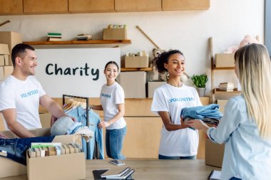 pretty african american girl giving clothes to woman while multicultural volunteers working in charity center clipart