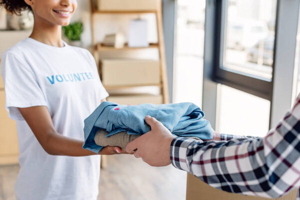 cropped view of african american volunteer giving clothes to man in charity center