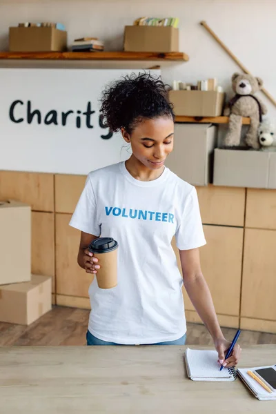 Bastante Afroamericano Voluntario Escribiendo Cuaderno Sosteniendo Taza Desechable — Foto de Stock