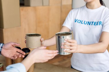 cropped view of volunteer giving canned food to man and woman in charity center clipart