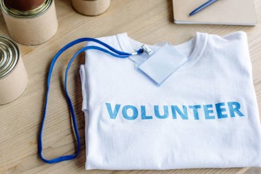 Mavi gönüllü yazılı beyaz tişört, rozet ve teneke kutu ahşap masa.