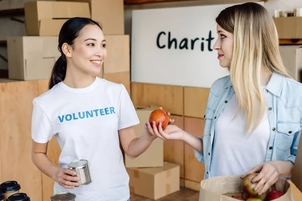 チャリティーセンターで女性に缶詰やリンゴを与える陽気なアジアのボランティア — ストック写真