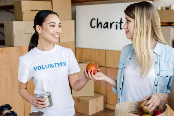 cheerful asian volunteer giving canned food and apples to woman in charity center
