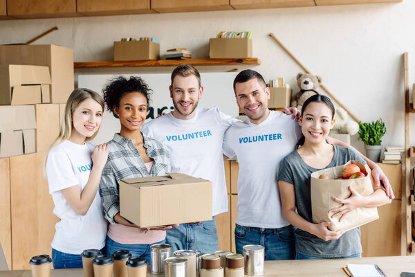 happy multicultural women holding cardboard box and paper bag with apples while standing near volunteers in charity center