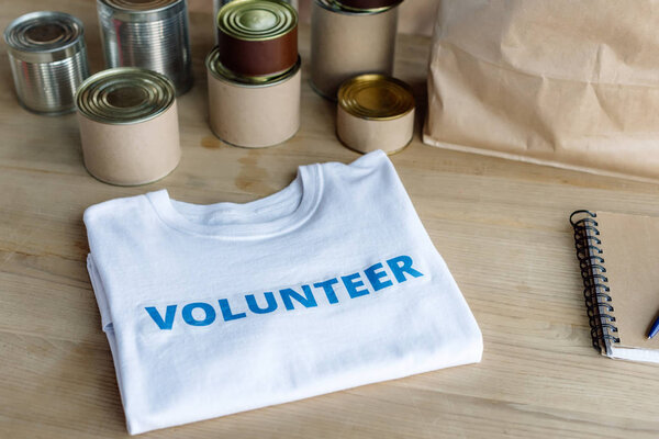 белая футболка с надписью добровольца, банками, бумажным пакетом и блокнотом на деревянном столе
