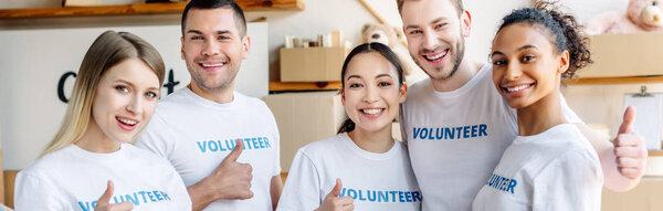 panoramic shot of young, cheerful volunteers showing thumbs up and looking at camera