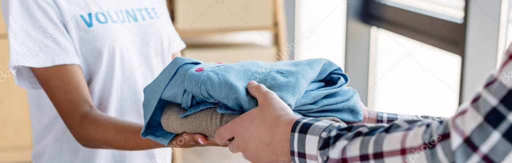 panoramic shot of african american volunteer giving clothes to man in charity center