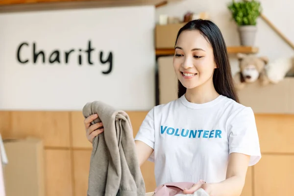 Mladý Usmívající Asijský Dobrovolník Drží Oblečení Zatímco Stojí Plakátu Charitativním — Stock fotografie