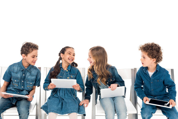 four smiling kids in denim clothes sitting on chairs and using digital tablets isolated on white
