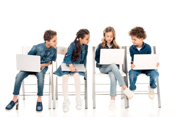 four kids in denim clothes sitting on chairs and using laptops on white