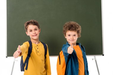 sırt çantaları kara tahta yakınında duran ve beyaz izole başparmak gösteren iki okul ların ön görünümü