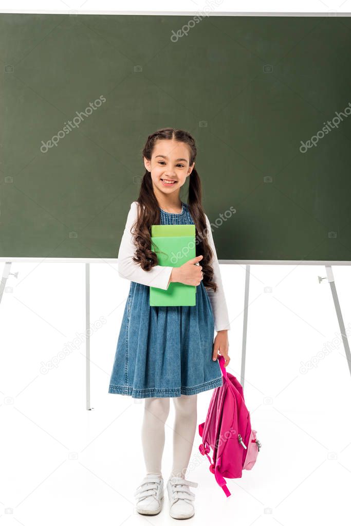 full length view of smiling schoolgirl with backpack and books standing near blackboard isolated on white