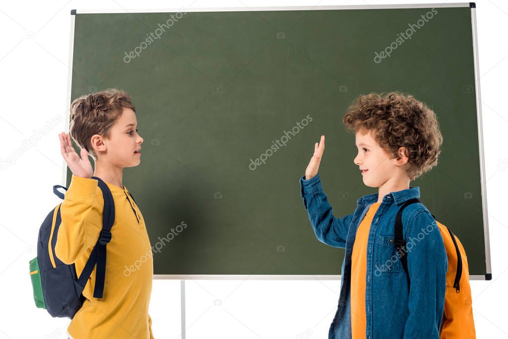two smiling schoolboys with backpacks showing high five sign near blackboard isolated on white