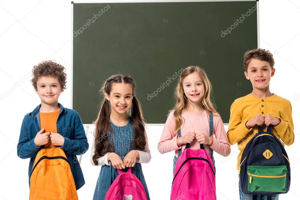 front view of smiling schoolchildren holding backpacks near blackboard isolated on white