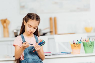 evde masanın yanında dururken renkli keçe kalem ve kalem tutan sevimli çocuk 
