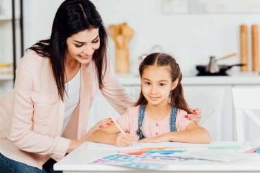 happy mother standing near paper near happy daughter looking at camera  clipart