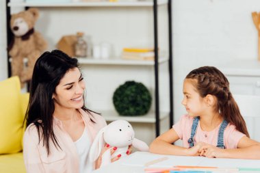 evde sevimli kızı yakın yumuşak oyuncak tutan neşeli anne 