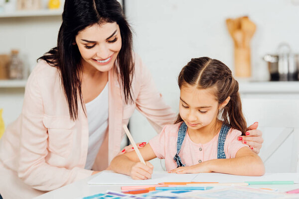 happy mother looking at cute daughter drawing on paper at home 