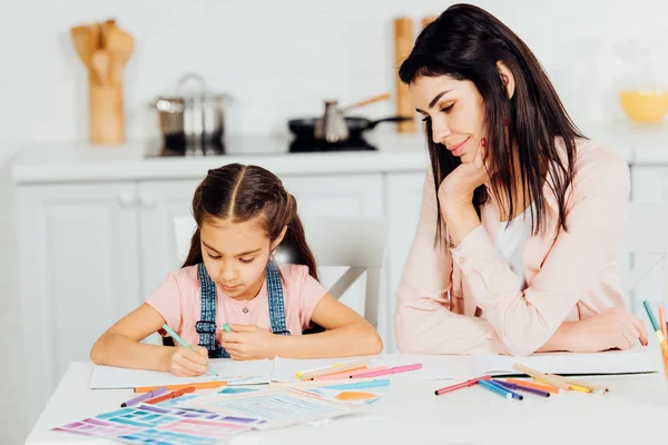 happy woman looking at daughter drawing on paper at home