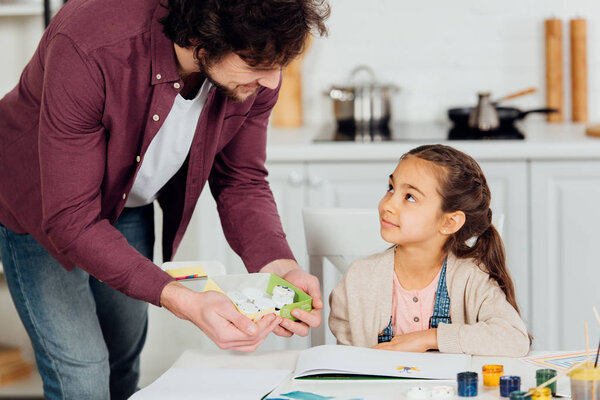 happy father holding box with gouache jars near cute daughter 