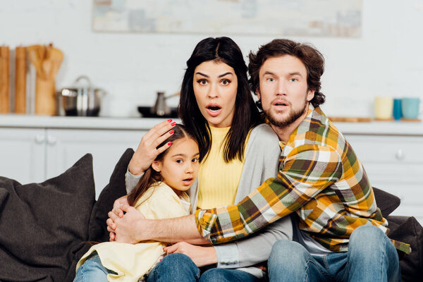shocked husband and wife watching tv and hugging cute daughter 