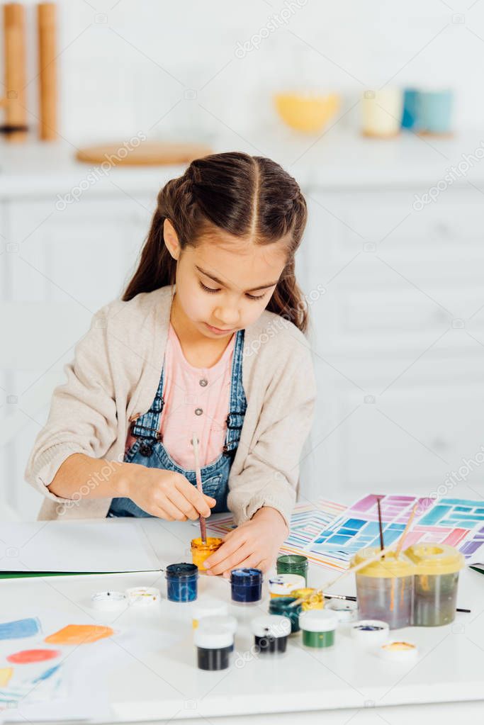 cute kid looking at yellow paint while holding paintbrush at home 