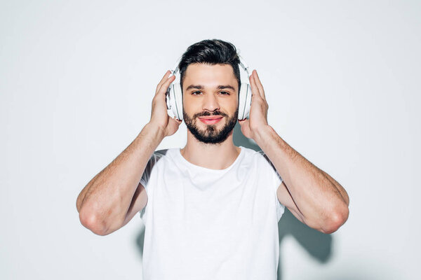 cheerful man touching headphones while listening music and smiling on white 