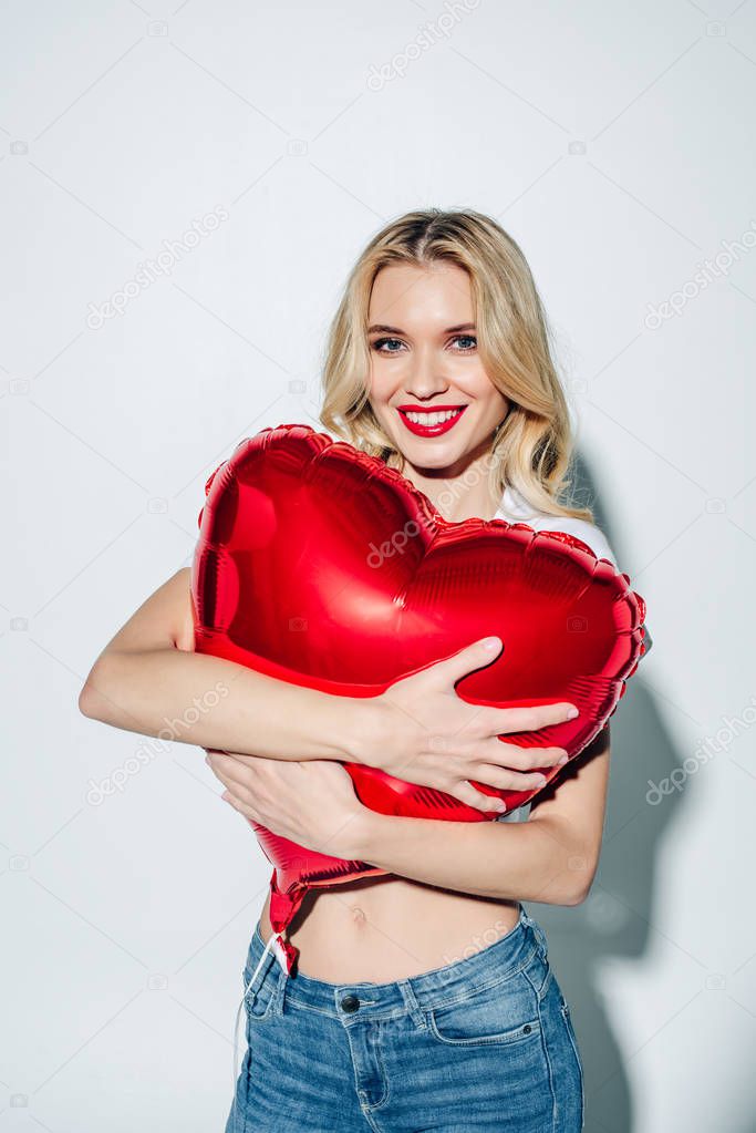 cheerful blonde girl hugging red heart-shaped balloon while standing on white 