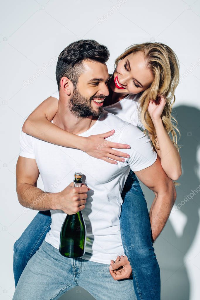 happy man holding bottle of champagne while piggybacking cheerful girl on white 