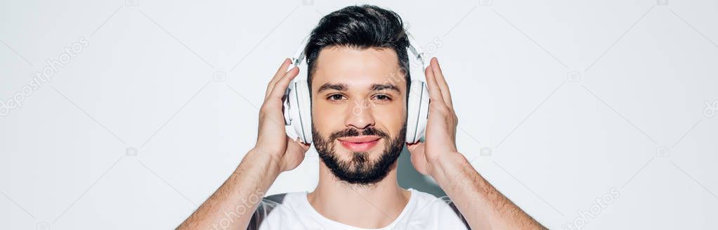 panoramic shot of cheerful man touching headphones while listening music and smiling on white 