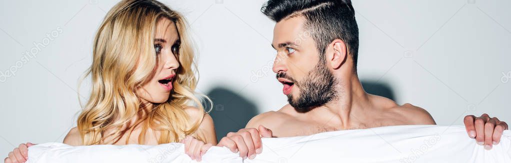 panoramic shot of surprised blonde woman and handsome man looking at each other while holding blanket on white 