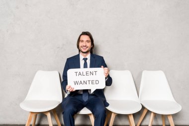 happy recruiter sitting on chair and holding speech bubble with talent wanted lettering  clipart