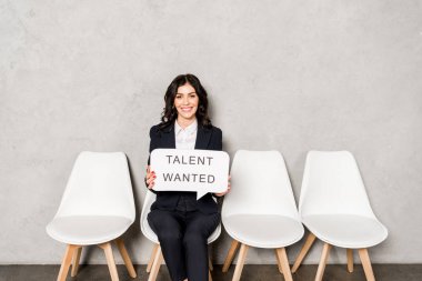 happy brunette woman holding speech bubble with talent wanted lettering while sitting on chair  clipart