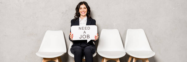 panoramic shot of happy brunette woman holding speech bubble with need a job lettering while sitting on chair 