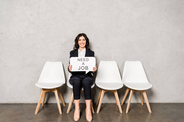 happy brunette woman holding speech bubble with need a job lettering while sitting on chair 