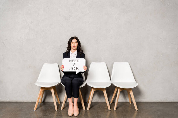 attractive brunette woman holding speech bubble with need a job lettering while sitting on chair 