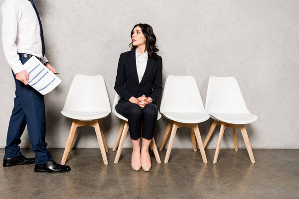 cropped view of man holding resume and walking near attractive brunette woman sitting on chair with clenched hands 