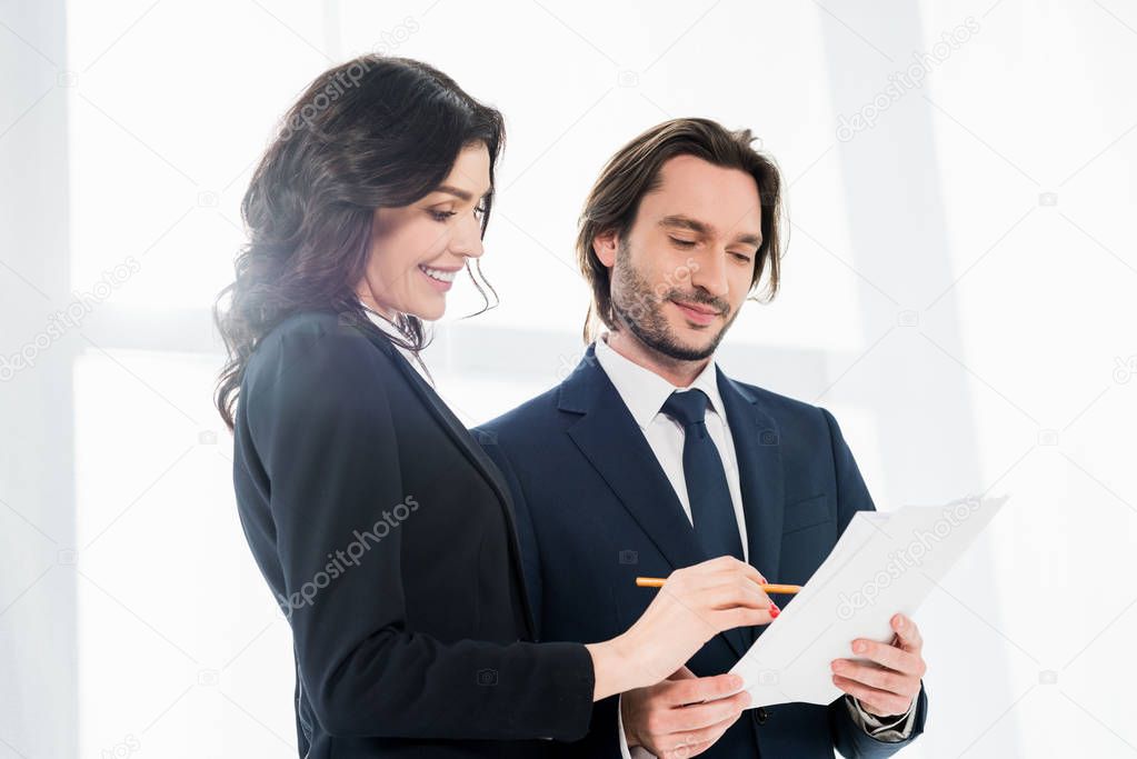 low angle view of cheerful woman standing near coworker and looking at paper 