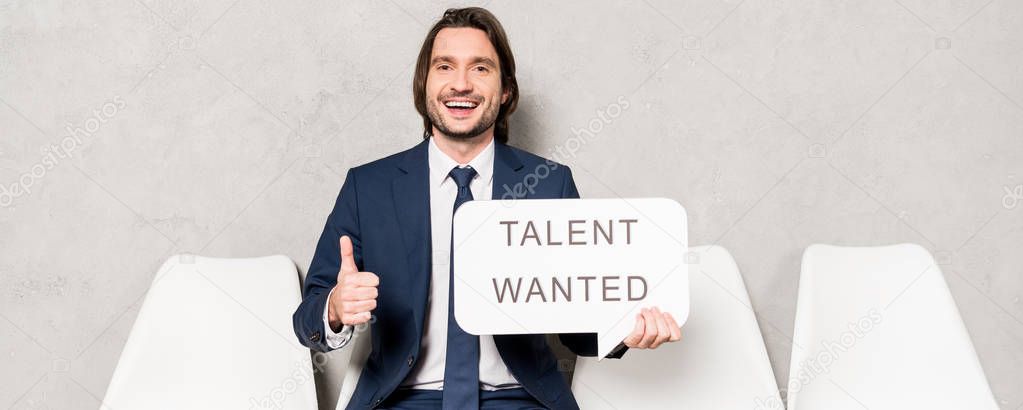 panoramic shot of happy recruiter sitting on chair and holding speech bubble with talent wanted lettering and showing thumb up