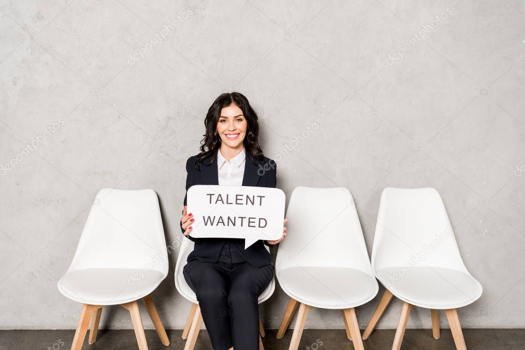 happy brunette woman holding speech bubble with talent wanted lettering while sitting on chair 