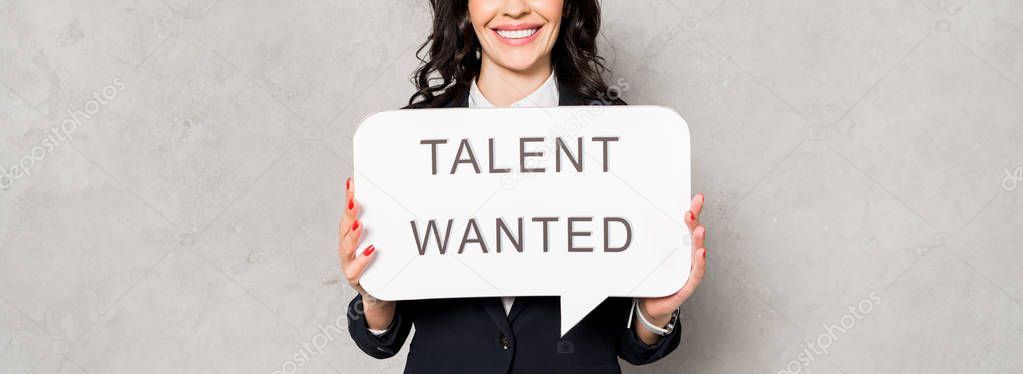 panoramic shot of happy brunette woman holding speech bubble with talent wanted lettering on grey 