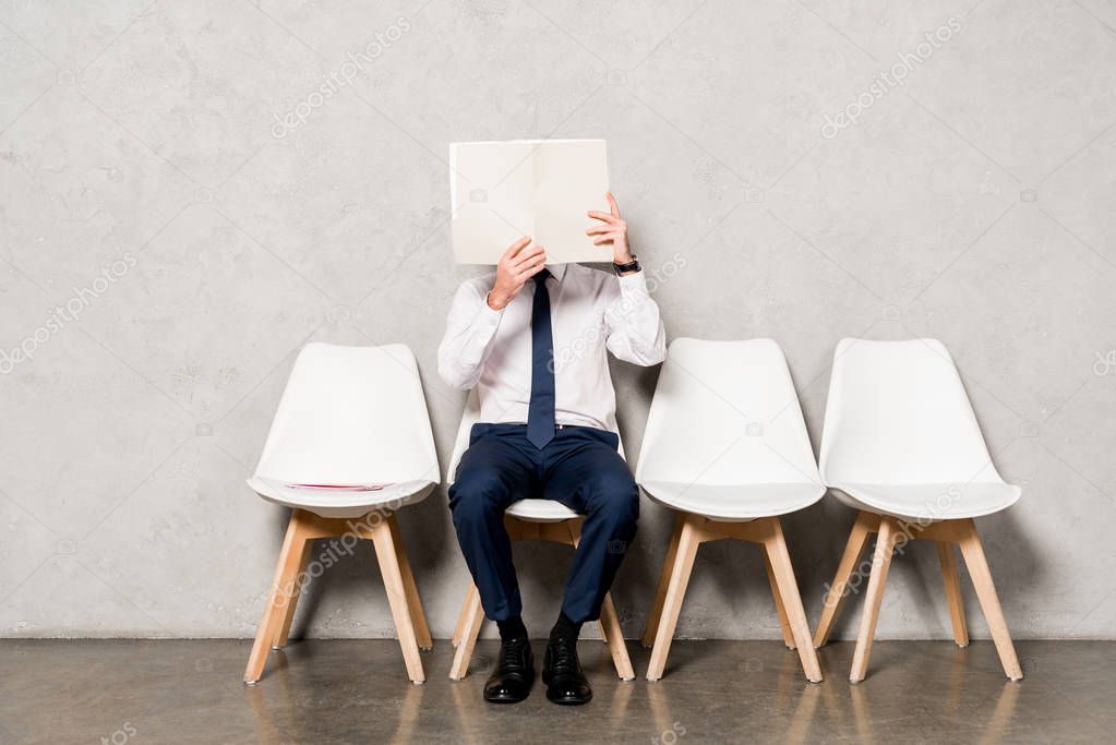 man in formal wear covering face with blank paper and sitting on chair 