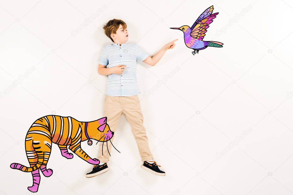top view of surprised kid pointing with finger humming bird near orange cat on white 