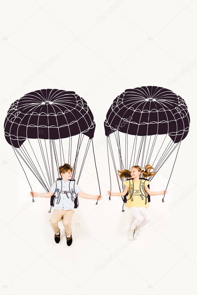 top view of cheerful boy and happy girl holding parachutes while flying on white 