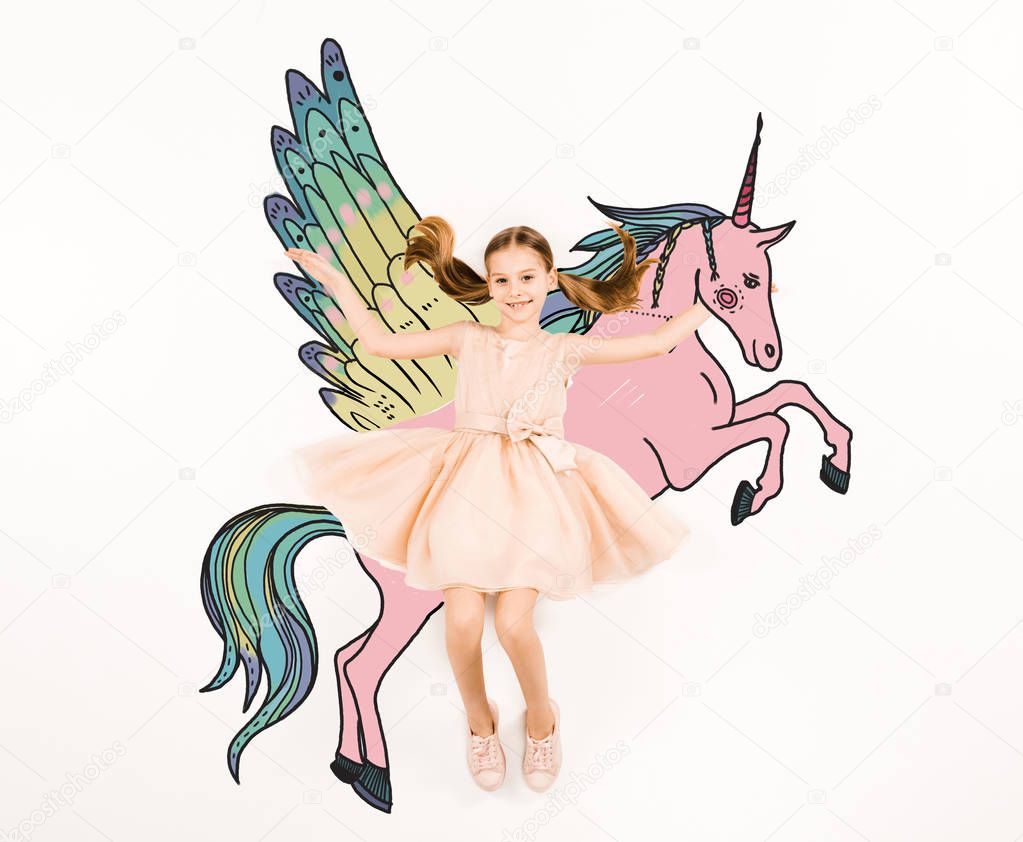 top view of cheerful kid looking at camera and gesturing near unicorn on white 