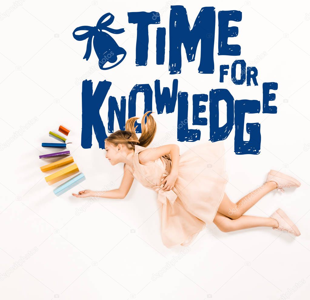 top view of cheerful kid in pink dress holding books and flying near time for knowledge lettering on white