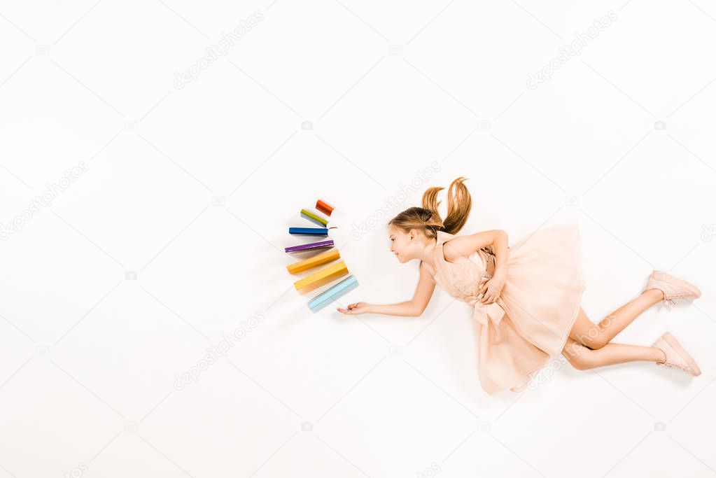 top view of cheerful kid in pink dress holding books and flying on white